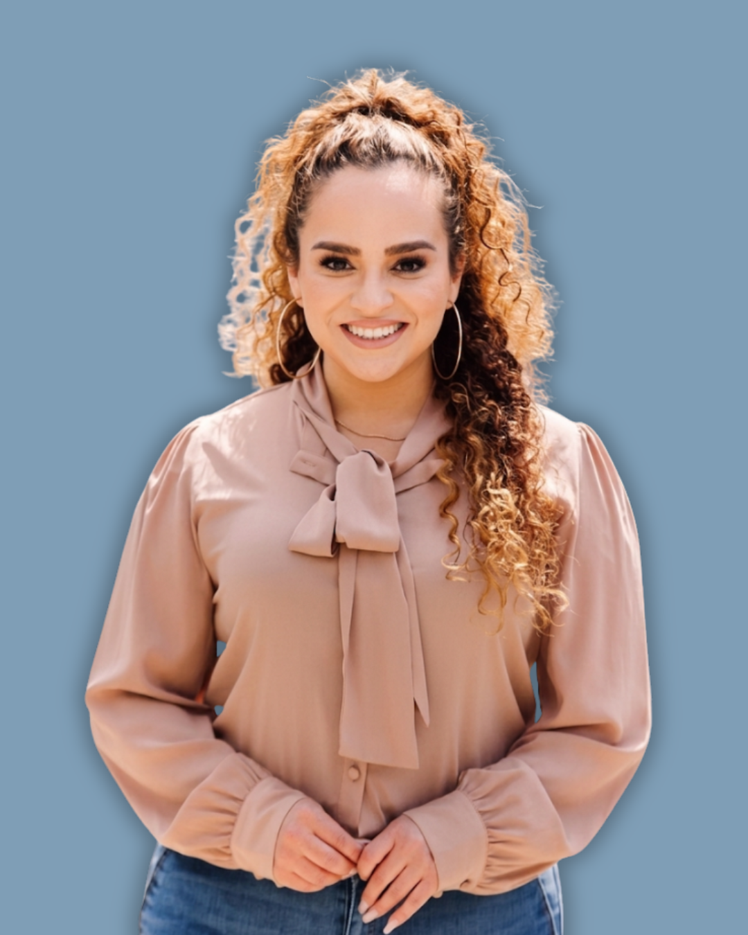 A professional studio portrait of a smiling female real estate agent with curly hair in a high ponytail, wearing a beige tie-neck blouse and jeans against a solid blue background.