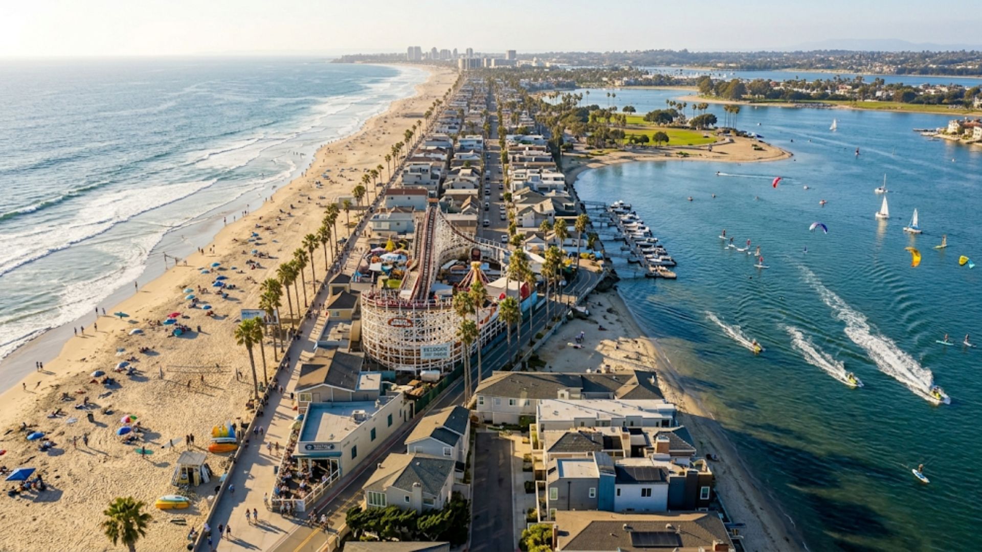 An aerial drone photograph of the Mission Beach peninsula in San Diego, showcasing the sandy beachfront on the Pacific Ocean, the calm waters of Mission Bay, residential housing, and the iconic Belmont Park roller coaster.