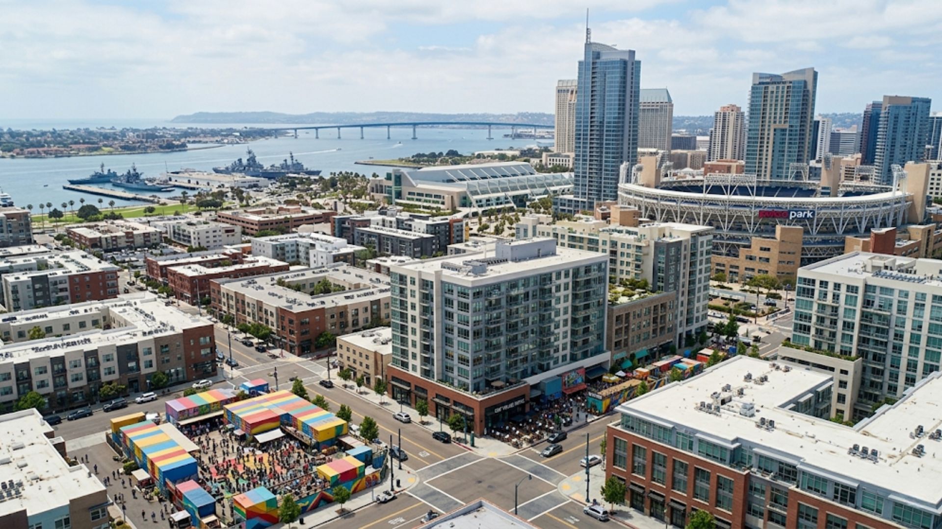 A high-angle aerial drone photograph overlooking the East Village neighborhood of San Diego, featuring Petco Park stadium, mid-rise and high-rise residential buildings, a colorful shipping container market, and the Coronado Bridge over the bay.