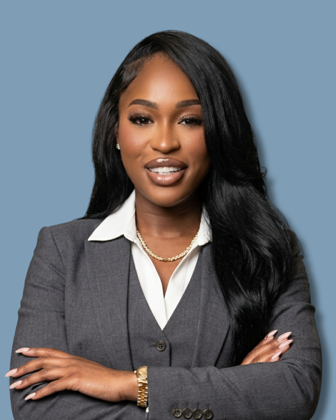 A professional corporate headshot of a smiling female real estate agent with long dark hair wearing a gray business suit and gold jewelry with crossed arms against a solid slate blue background